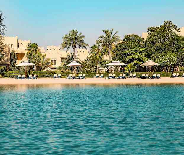 Udsigt over en strand med liggestole og parasoller, omgivet af palmer og bygninger med traditionel arkitektur i baggrunden. (AI-genereret tekst)