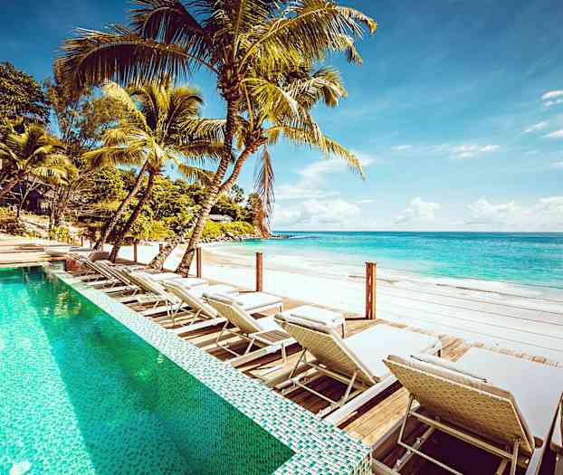 Udendørs pool med liggestole ved en sandstrand med klart blåt vand. (AI-genereret tekst)