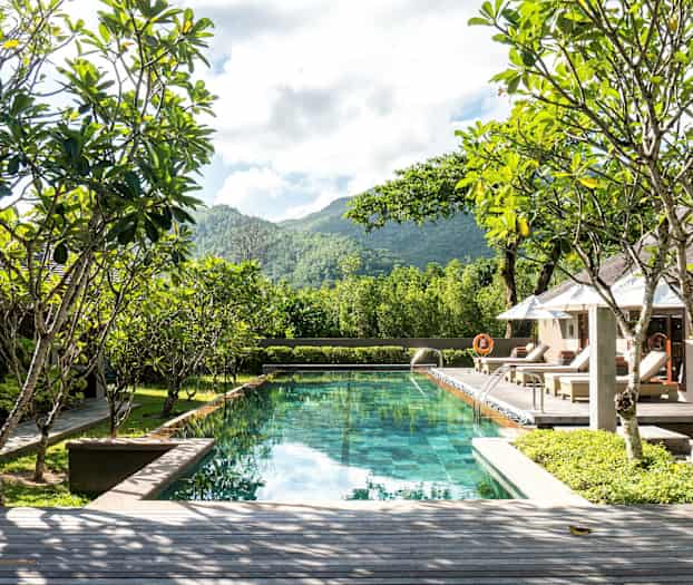 Billede af en swimmingpool omgivet af grønne områder og liggestole, med udsigt til bakker og træer. (AI-genereret tekst)