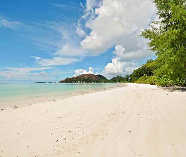 En sandstrand med klart vand og grøn vegetation i baggrunden. En bjergtop er synlig i horisonten. (AI-genereret tekst)