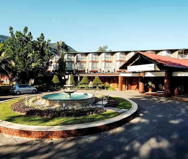 Hotelkompleks med store vinduer, velholdt have og en fontæne, beliggende mod en baggrund af bjerge. (AI-genereret tekst)