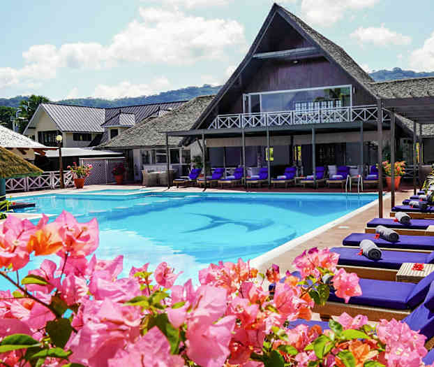 Poolområde med liggestole og blomster, bygninger i baggrunden. (AI-genereret tekst)