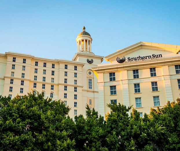 En lys bygning med flere etager og et tårn kan ses bag frodige træer. (AI-genereret tekst)