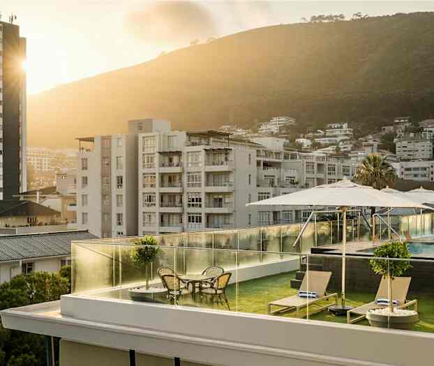 Tagterrasse med liggestole, borde og parasoller, swimmingpool i baggrunden samt et bymiljø med frodige bakker. (AI-genereret tekst)