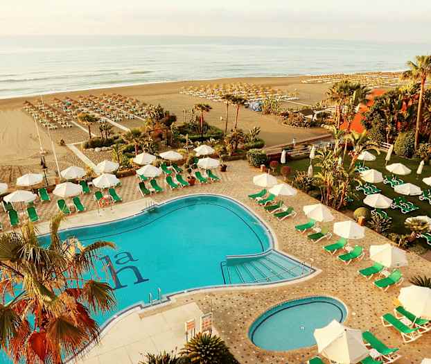 En udendørs pool med omkringliggende liggestole og parasoller, strand synlig i baggrunden. (AI-genereret tekst)