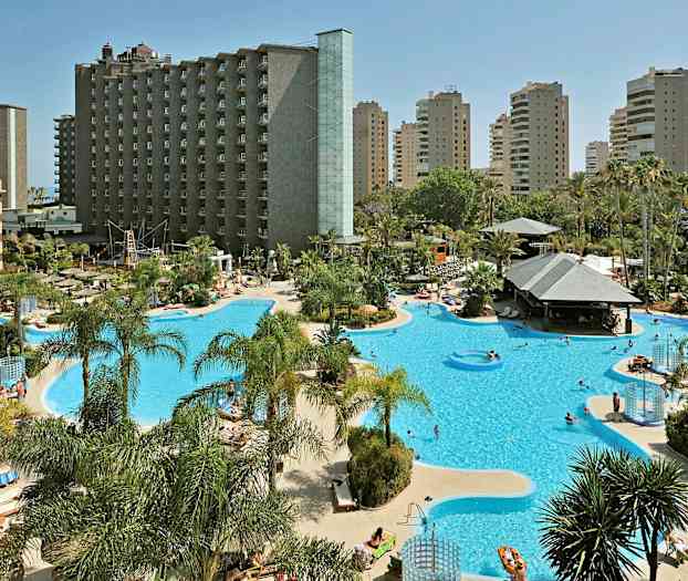 Et hotelområde med stor pool og liggestole, omgivet af palmer og højhuse. (AI-genereret tekst)