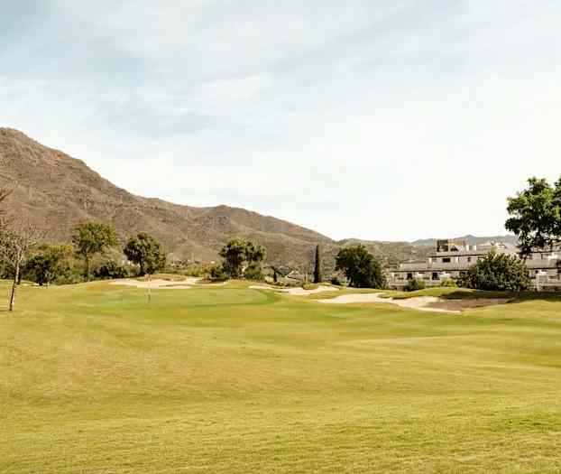 En golfbane omgivet af grøn vegetation, med bygninger og bjerge i baggrunden. (AI-genereret tekst)