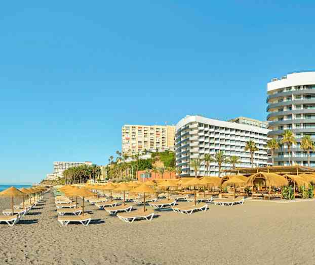 En strand med liggestole og parasoller, omgivet af flere hotelbygninger. (AI-genereret tekst)