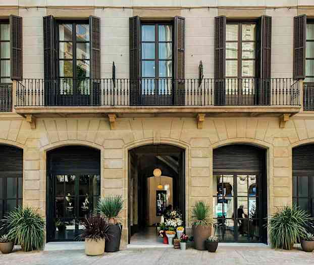 Billede af facaden på en bygning med flere høje vinduer og en indgang med dekorationer af blomsterpotter. (AI-genereret tekst)