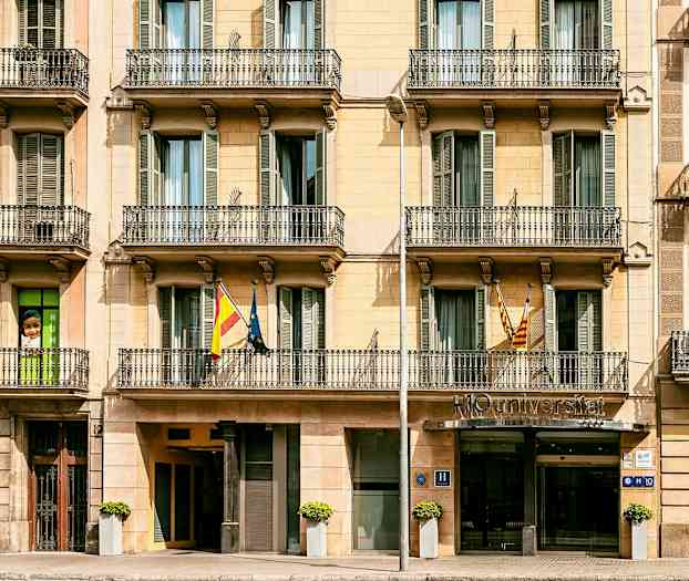 Forsiden af en bygning med altaner, indgang til venstre og et hotelskilt ved indgangen. (AI-genereret tekst)