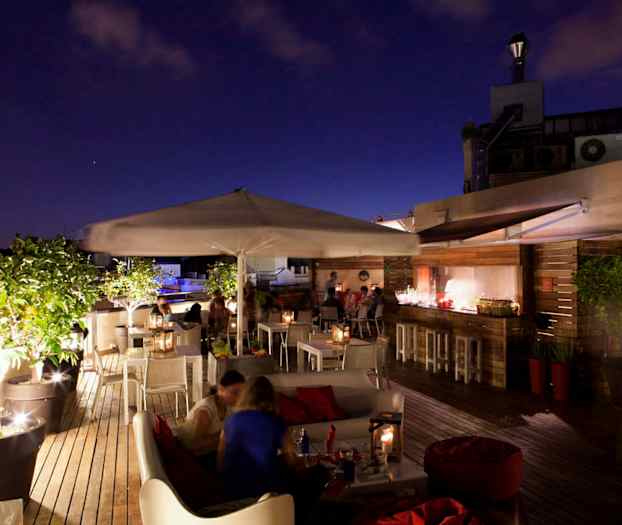 En tagterrasse med siddepladser, borde, parasoller og grønne planter, oplyst i aftenomgivelser. (AI-genereret tekst)