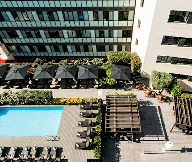 Luftfoto af en pool med liggestole, parasoller og omkringliggende bygninger. (AI-genereret tekst)