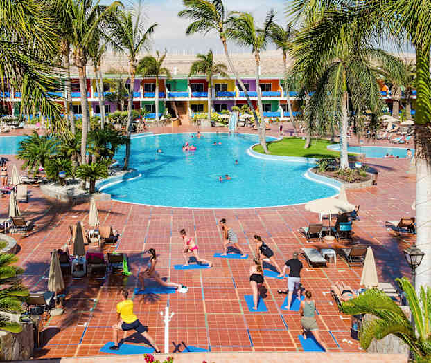 Ved et stort hotel med farverige bygninger dyrker folk udendørs yoga på måtter ved siden af en pool. (AI-genereret tekst)