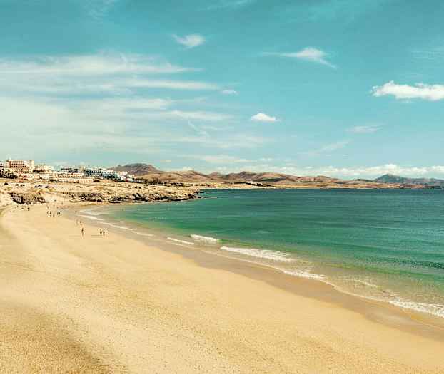 En bred sandstrand, der strækker sig langs kysten med turkisblåt vand og bygninger i baggrunden. (AI-genereret tekst)