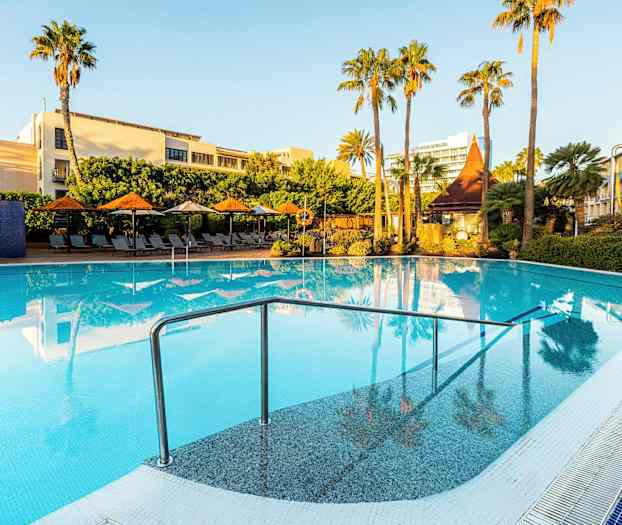 En udendørs swimmingpool omgivet af liggestole, parasoller og grønne palmer under en klar blå himmel. (AI-genereret tekst)