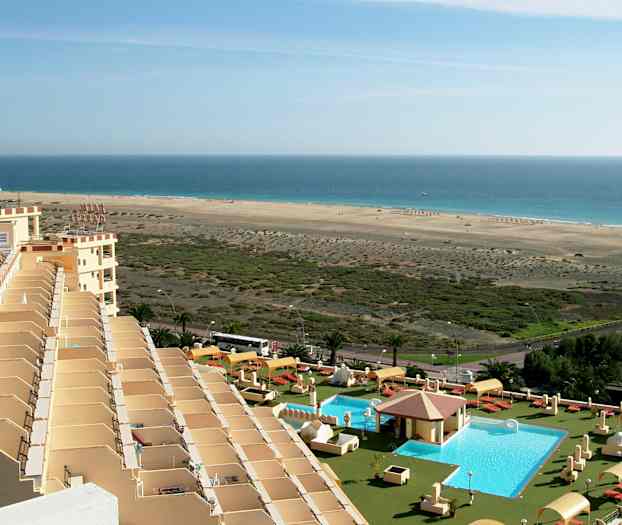 Billede af et hotelområde med en pool, liggestole og parasoller i forgrunden, og en lang sandstrand ved havet i baggrunden. (AI-genereret tekst)