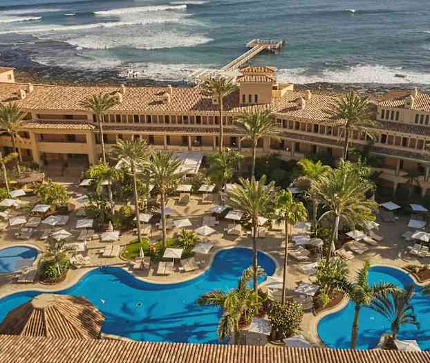 Hotelområde med swimmingpools, liggestole og parasoller omgivet af palmer. I baggrunden ses havet med bølger og en bro. (AI-genereret tekst)