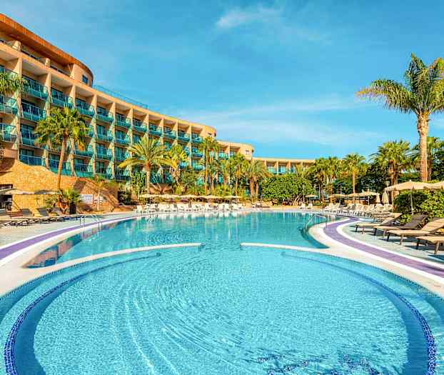 Hotellets poolområde med liggestole og palmer foran en bygning med altaner. (AI-genereret tekst)