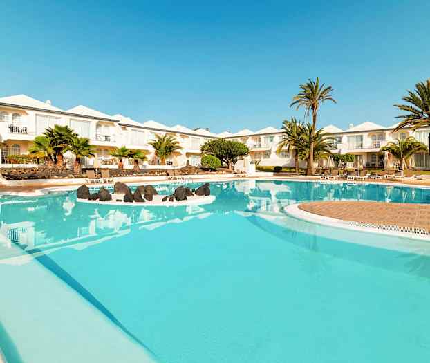 Et billede af en hotelpool med klart blåt vand, omgivet af palmetræer og hvide bygninger i baggrunden. (AI-genereret tekst)