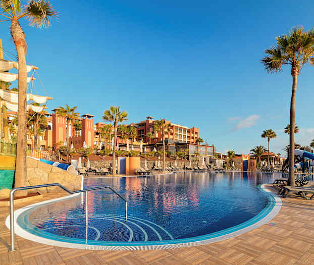 En udendørspool med soldæk, omgivet af palmetræer og med en bygning i baggrunden. I forgrunden ses et skib som udsmykning. (AI-genereret tekst)