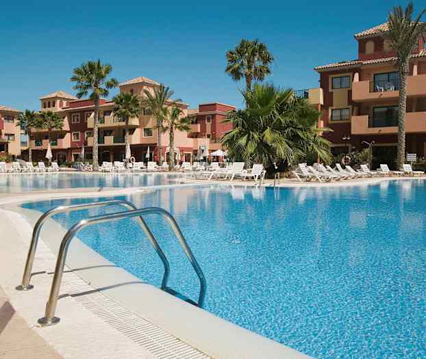 Et feriekompleks med en udendørs pool omgivet af solsenge og palmer, samt bygninger i baggrunden. (AI-genereret tekst)