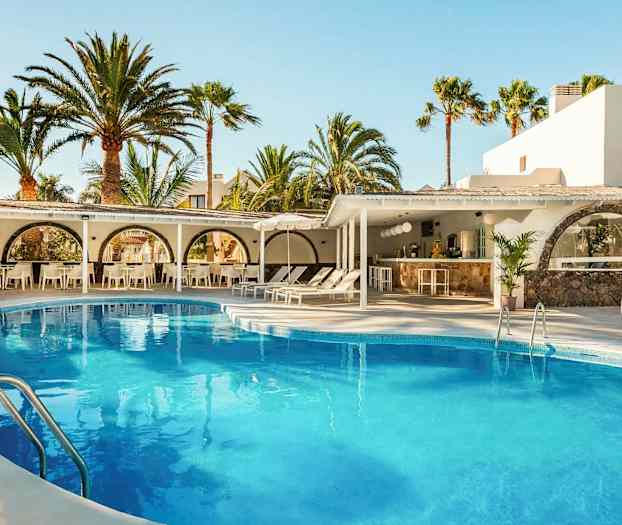 En udendørs swimmingpool omgivet af liggestole og parasoller, med hvide bygninger og høje palmer i baggrunden. (AI-genereret tekst)