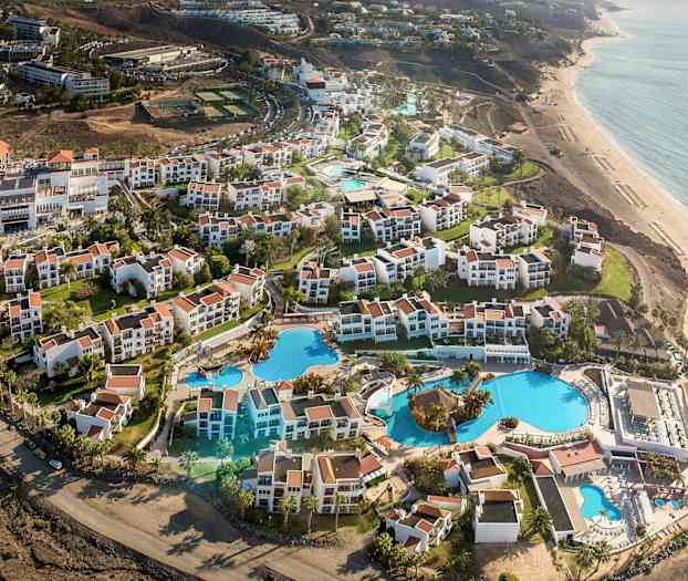 Luftfoto af et feriested med hvide bygninger og røde tage, samt store swimmingpools og haver nær stranden. (AI-genereret tekst)
