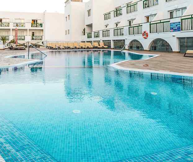 Udendørs swimmingpool på et feriested med liggestole og hvide bygninger i baggrunden. (AI-genereret tekst)