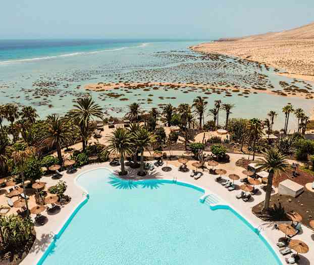 En pool omgivet af liggestole og parasoller med palmer i baggrunden samt udsigt over en lavvandet turkisblå strand og hav. (AI-genereret tekst)