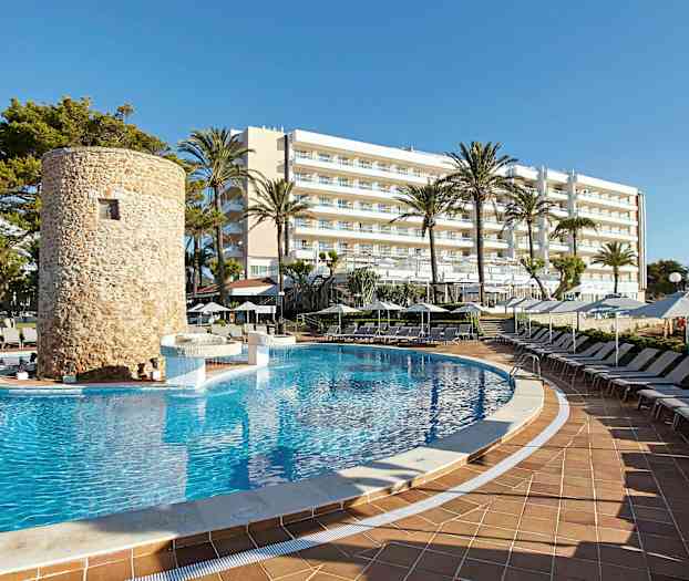 En swimmingpool omgivet af liggestole, med et stenbygget tårn og et hotel i baggrunden. (AI-genereret tekst)