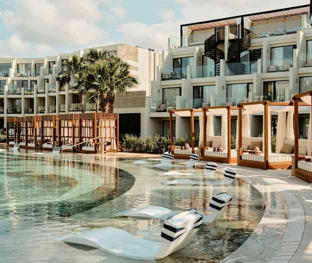 En hotelpool med liggestole i poolen og cabanas ved siden af. Bygningen har altaner med glasrækværk. (AI-genereret tekst)
