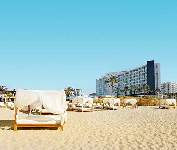 Strand med senge med hvide draperinger, i baggrunden ses en moderne bygning. (AI-genereret tekst)