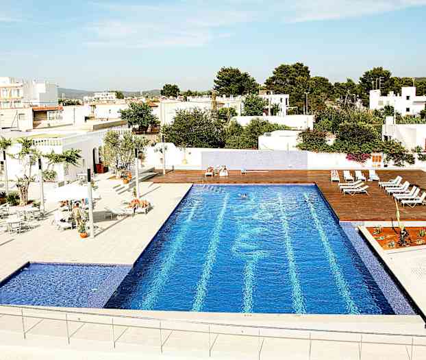 En udendørs pool med blå fliser, omgivet af liggestole og hvide bygninger. (AI-genereret tekst)