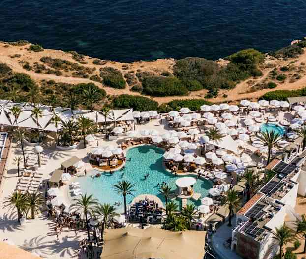 Luftfoto af en ferieresort med flere pools og hvide parasoller, ved siden af klipper og havet. (AI-genereret tekst)