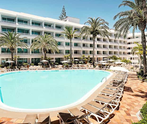 En hotelpool med solsenge og parasoller omgivet af frodige palmer, samt en fleretagers bygning med altaner i baggrunden. (AI-genereret tekst)