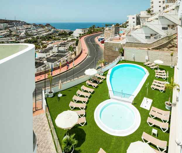 Udendørs pool med liggestole på grøn græsplæne og udsigt til nærliggende bygninger og havet. (AI-genereret tekst)