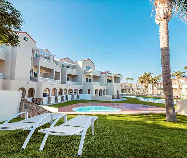 Udendørsområde med liggestole, græsplæne, pool samt nærliggende bygning i lyse farver. (AI-genereret tekst)