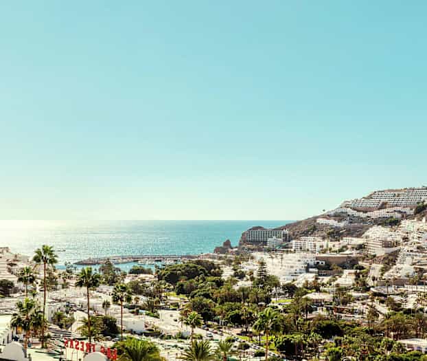 Udsigt over en kystby med hvide bygninger og palmer foran et glitrende hav. (AI-genereret tekst)