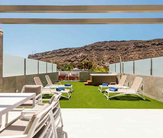 En udendørsterrasse med liggestole på kunstgræs og et spabad, omgivet af glasbarrierer og bjerge i baggrunden. (AI-genereret tekst)