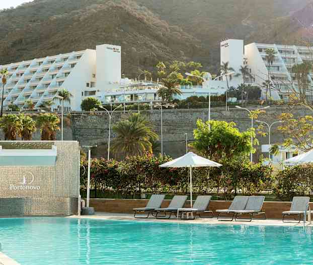 En pool med liggestole og parasoller, med et hvidt feriekompleks og bjerge i baggrunden. (AI-genereret tekst)