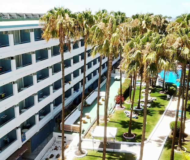 Hotel med hvide facader omgivet af græsplæne med palmer og en pool. (AI-genereret tekst)