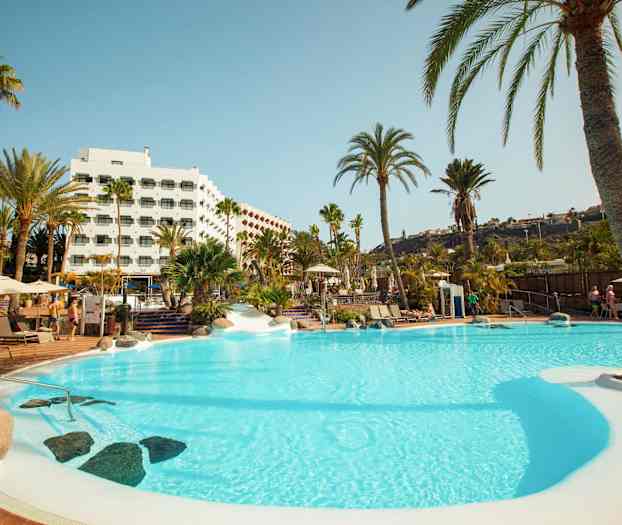 En swimmingpool omgivet af palmer og et hotel i baggrunden. (AI-genereret tekst)