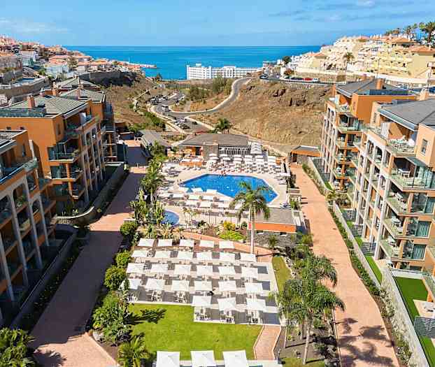 Hotelområde med pool, liggestole og omkringliggende bygninger, med havet i baggrunden. (AI-genereret tekst)