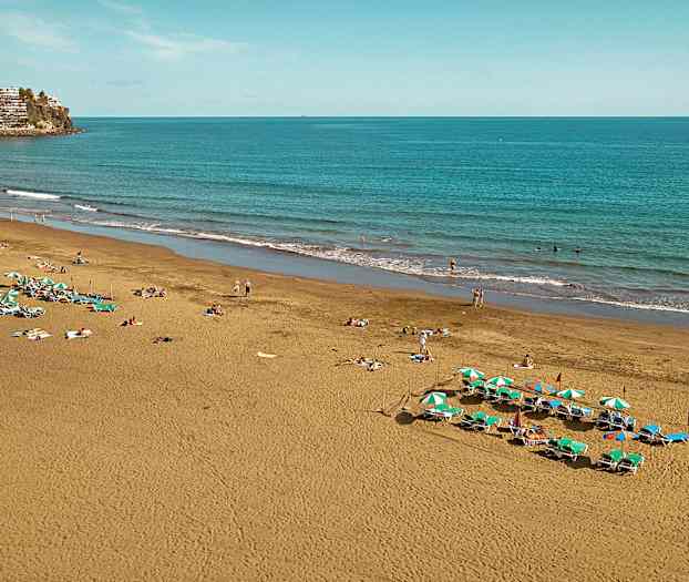 Billedet viser en sandstrand ved havet med liggestole og parasoller samt mennesker, der nyder stranden. (AI-genereret tekst)