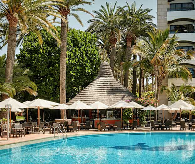 Udendørs område med parasoller foran en pool og en bygning med palmer i baggrunden. (AI-genereret tekst)