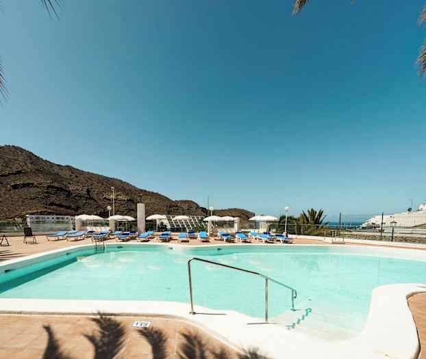 Udendørs pool med liggestole og parasoller, bjerge i baggrunden og klar blå himmel. (AI-genereret tekst)