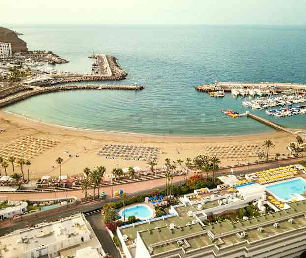 Luftfoto af en strand med parasoller og liggestole, en omkringliggende marina med både og nærliggende bygninger. (AI-genereret tekst)