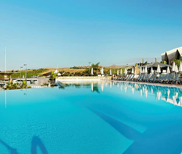 Stor udendørs pool med liggestole og parasoller i baggrunden, omgivet af beplantning og bygninger. (AI-genereret tekst)