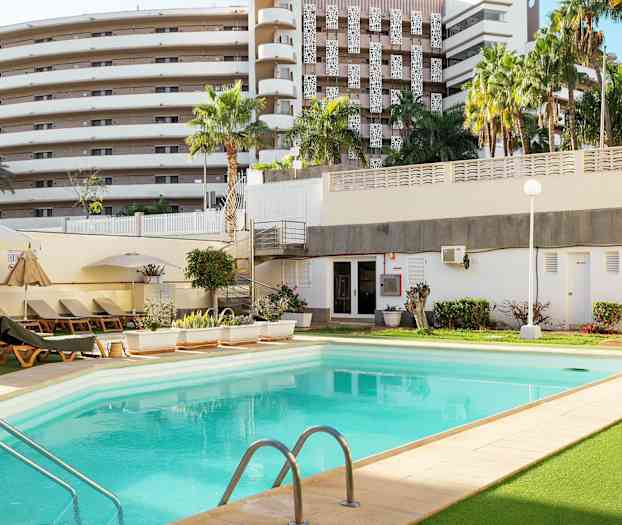 En udendørs swimmingpool omgivet af liggestole, beplantning og en bygning i baggrunden. (AI-genereret tekst)