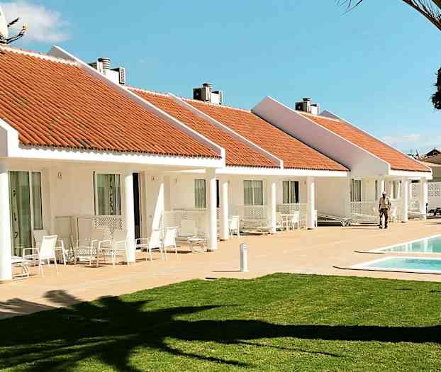 Hvide bungalower med orange tage og en pool foran. En græsplæne i forgrunden. (AI-genereret tekst)